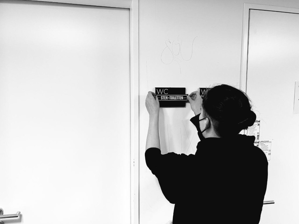 Eine leicht vermummte Person bringt einen Sticker auf einem Toiletten Schild an. Auf diesem Sticker steht: Steh-Toiletten.